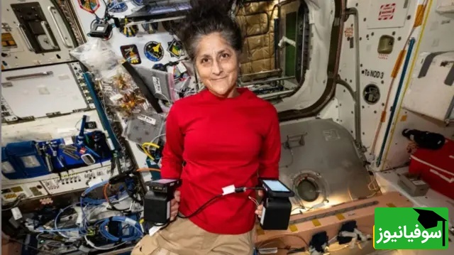 تصویر ویلیامز در داخل ایستگاه فضایی. او لباس سرخ بر تن دارد و در هر دو دستش تجهیزات را گرفته است. 